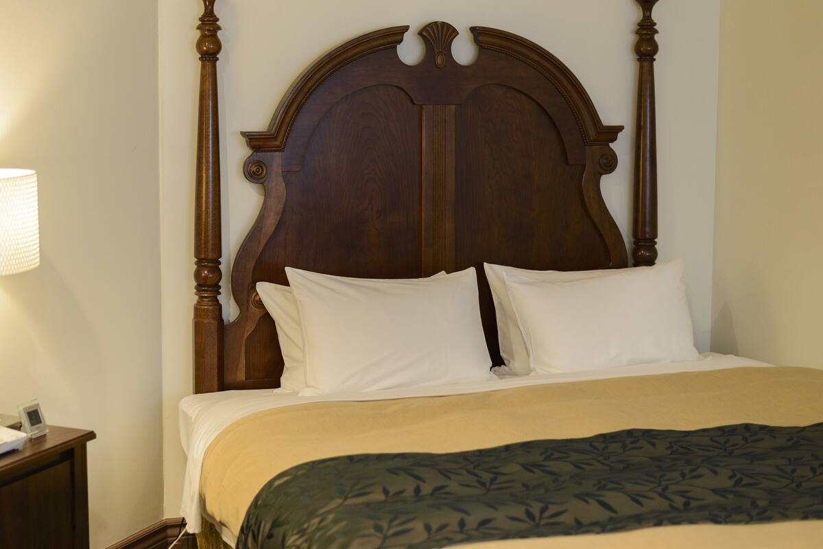 A neatly made bed with a wooden headboard, a bedside lamp, and a nightstand with a phone and a clock.