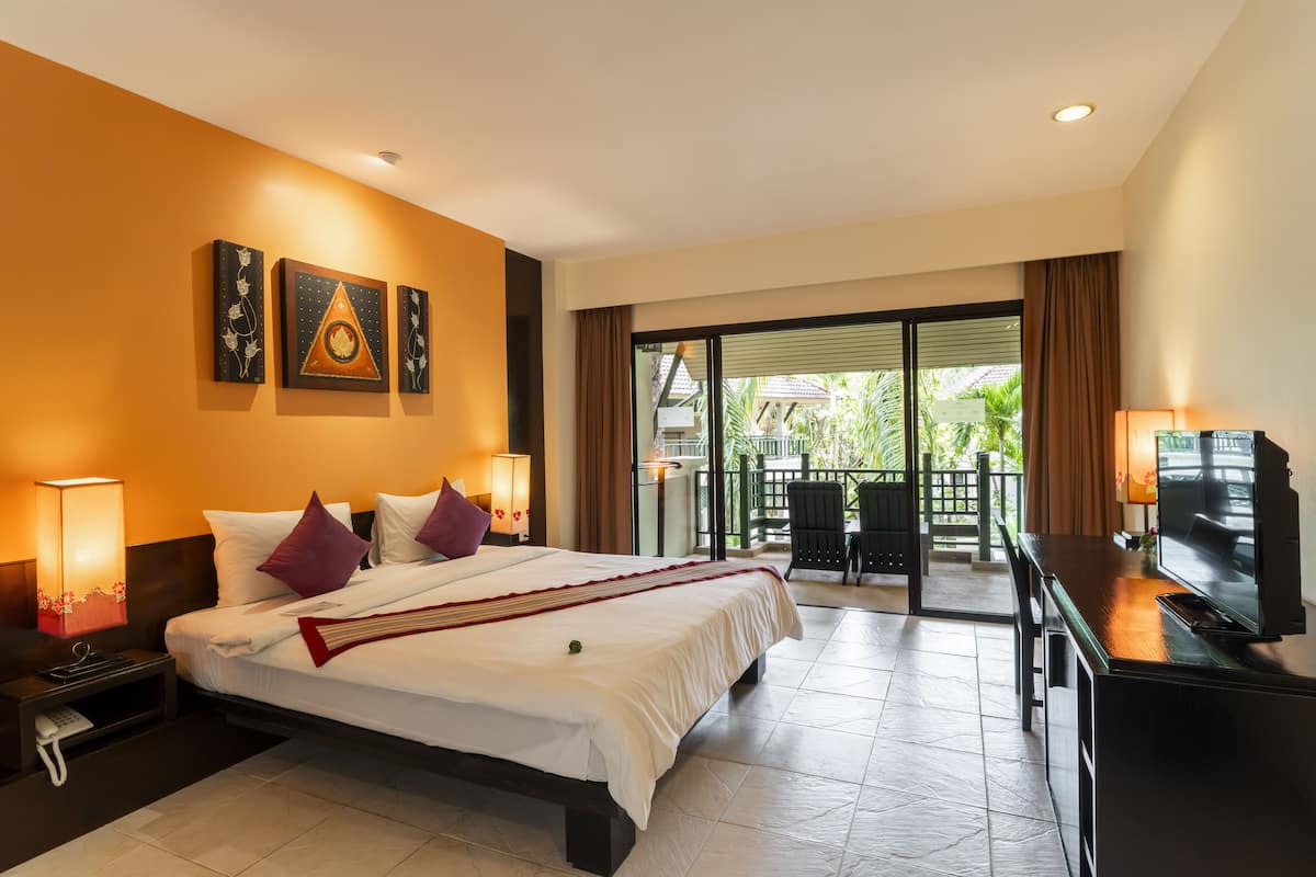 A hotel room with a bed, bedside lamps, a TV, and a balcony with a view of greenery.