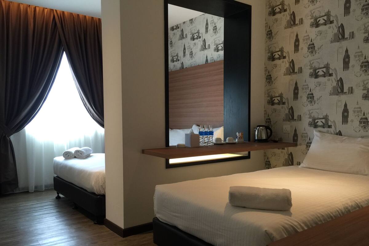 A hotel room with two beds, a large mirror, a wooden shelf with toiletries, and a window with curtains.