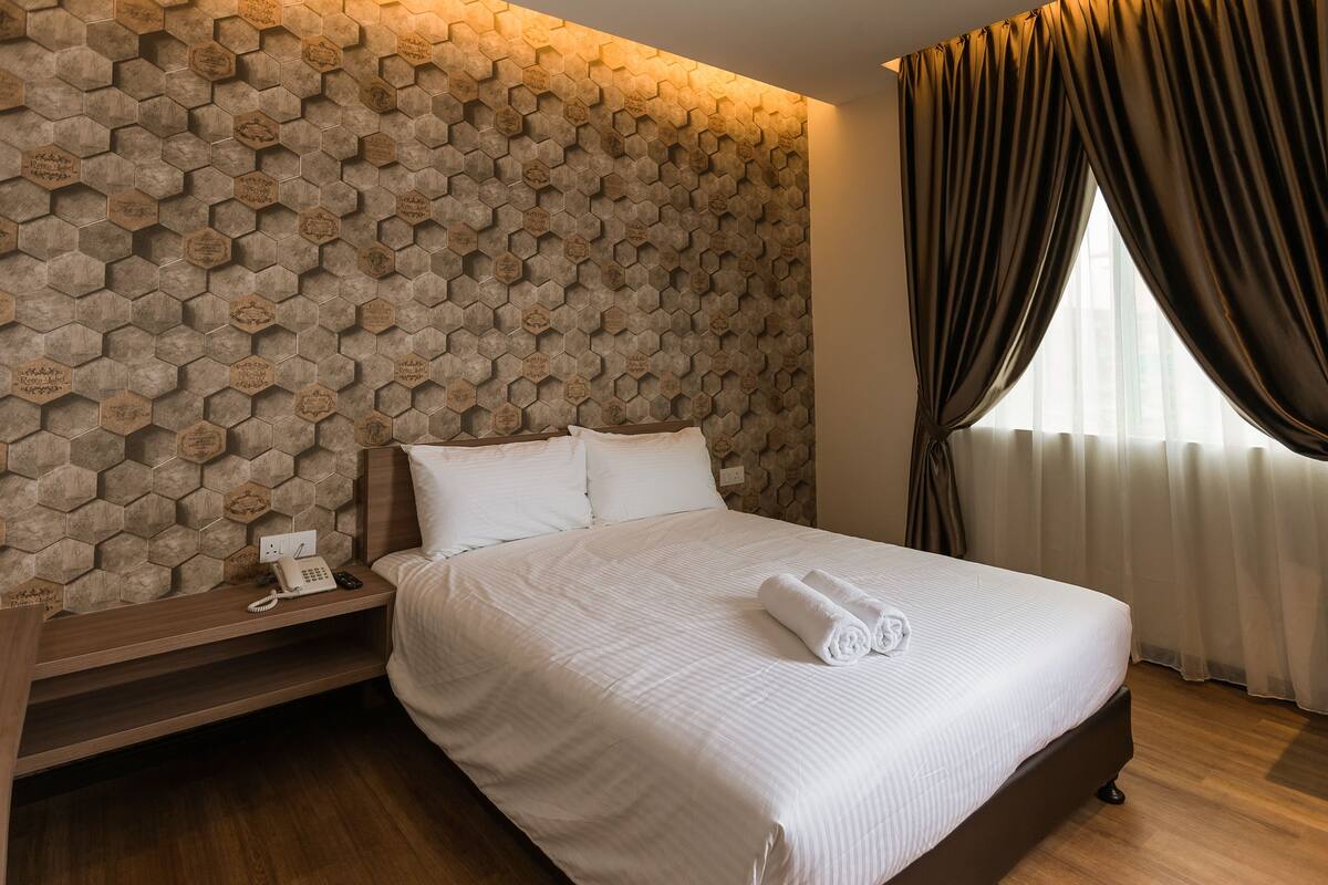 A hotel room with a large bed, a wooden bedside table with a phone, a wall with hexagonal patterns, and a window with curtains.
