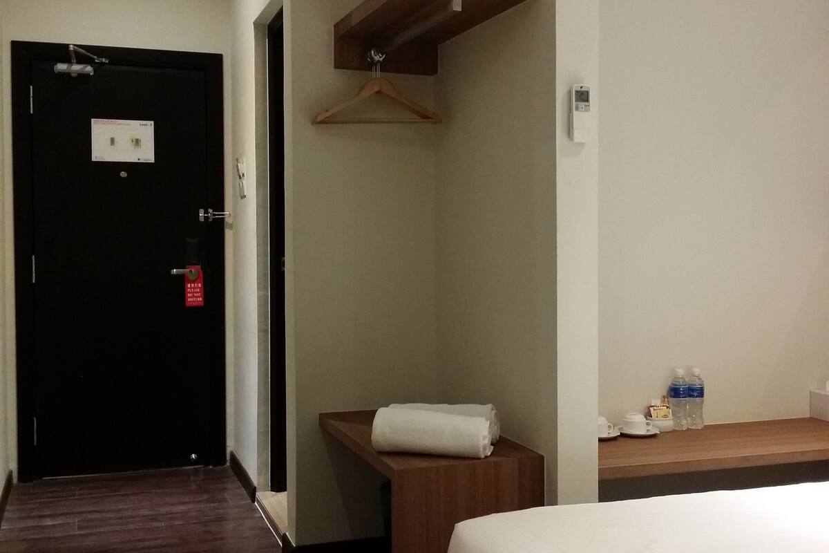 A hotel room with a bed, a wooden bedside table, a towel, a water bottle, and an air conditioning unit.