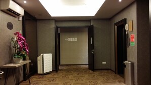 Un pasillo de hotel con alfombra estampada, una unidad de aire acondicionado, una planta en maceta, un refrigerador y un cubo de basura.