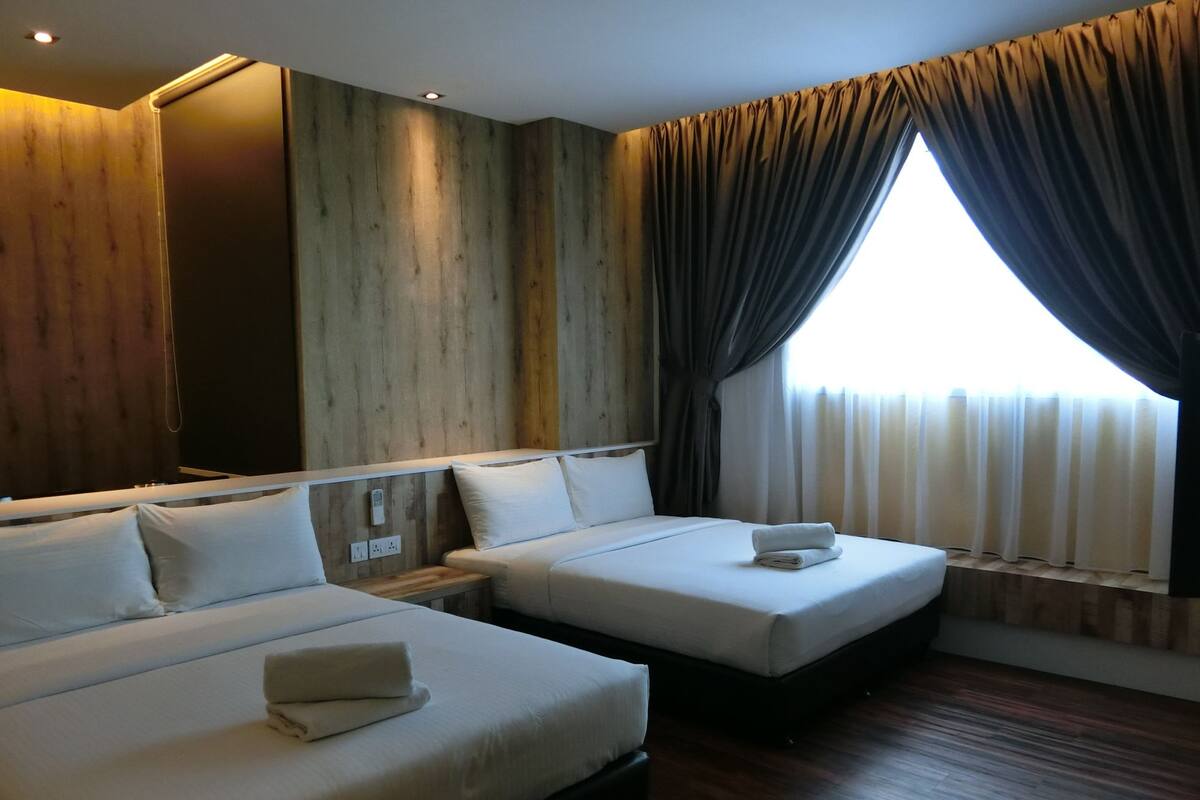 A hotel room with two beds, wooden paneling on the walls, a large window with curtains, and a bedside table with a lamp.