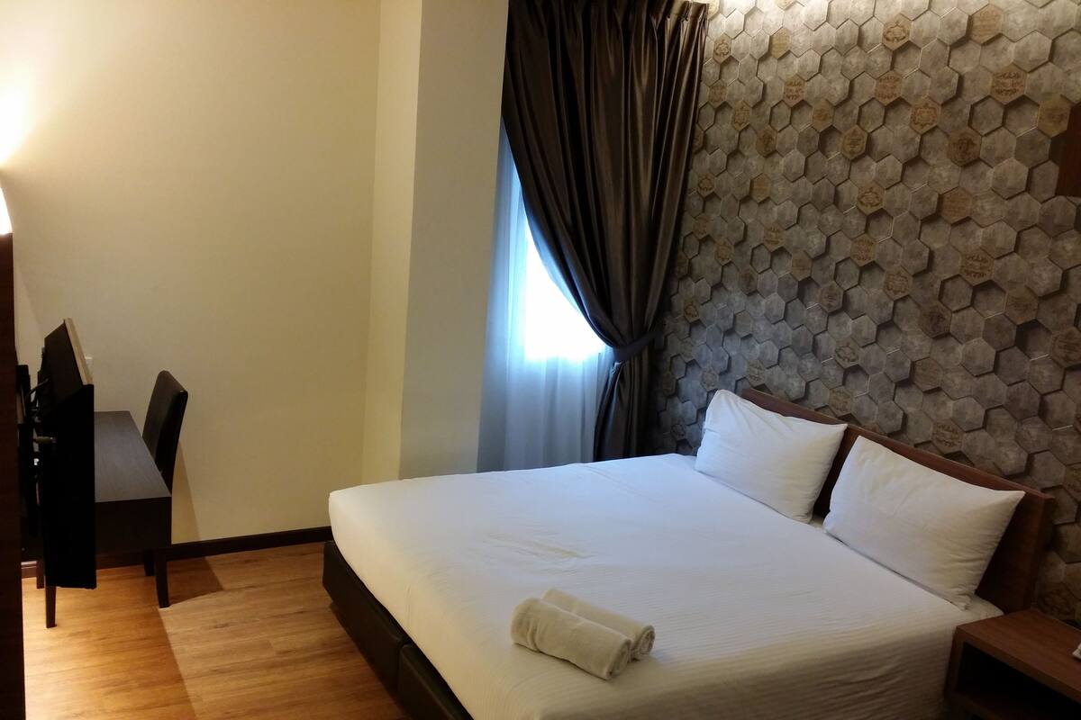 A hotel room with a large bed, a wooden headboard, a desk with a chair, and a window with curtains.