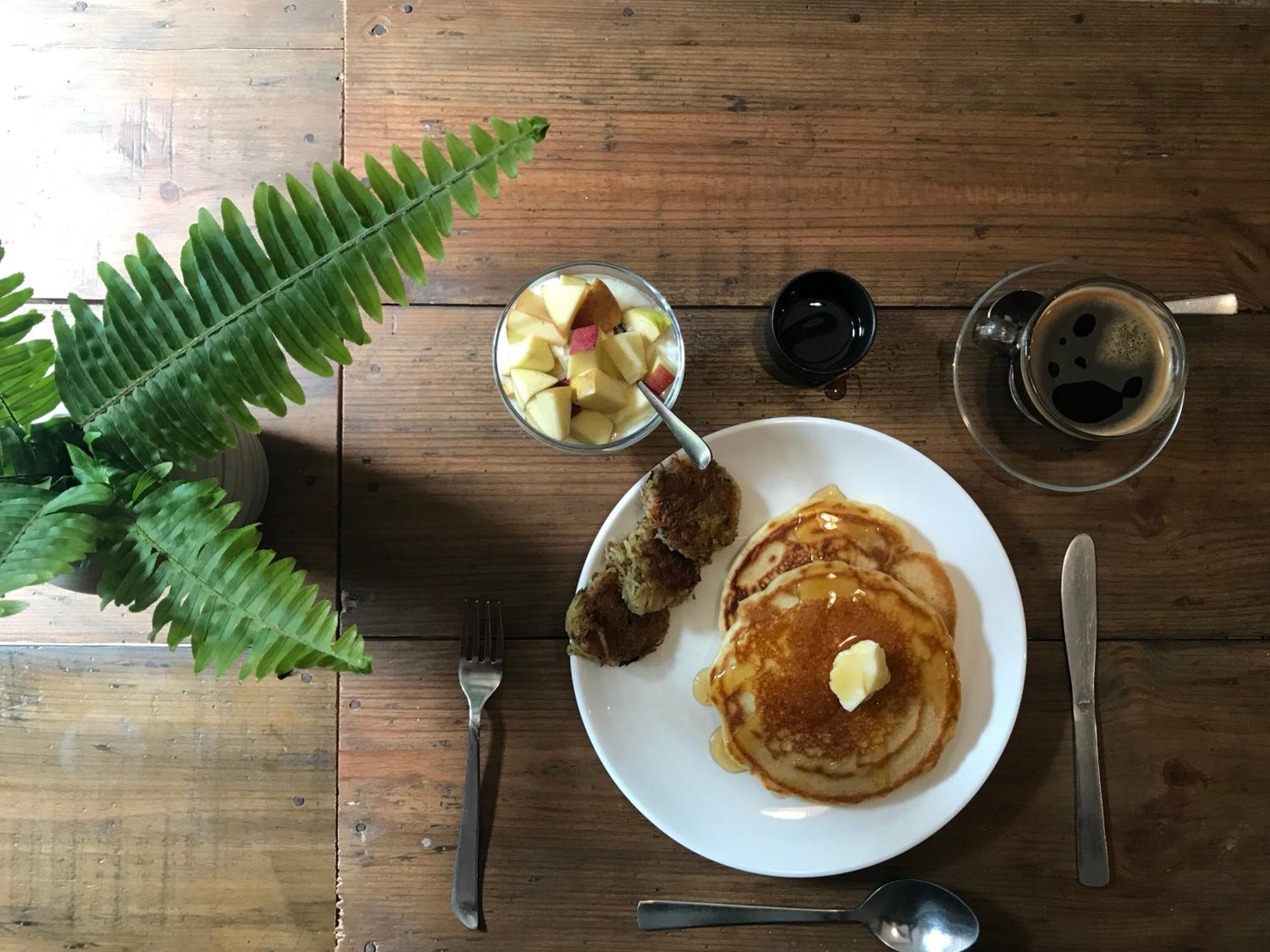 Una mesa de madera con una fuente de panqueques, un bol de fruta, una taza de café y un tenedor.