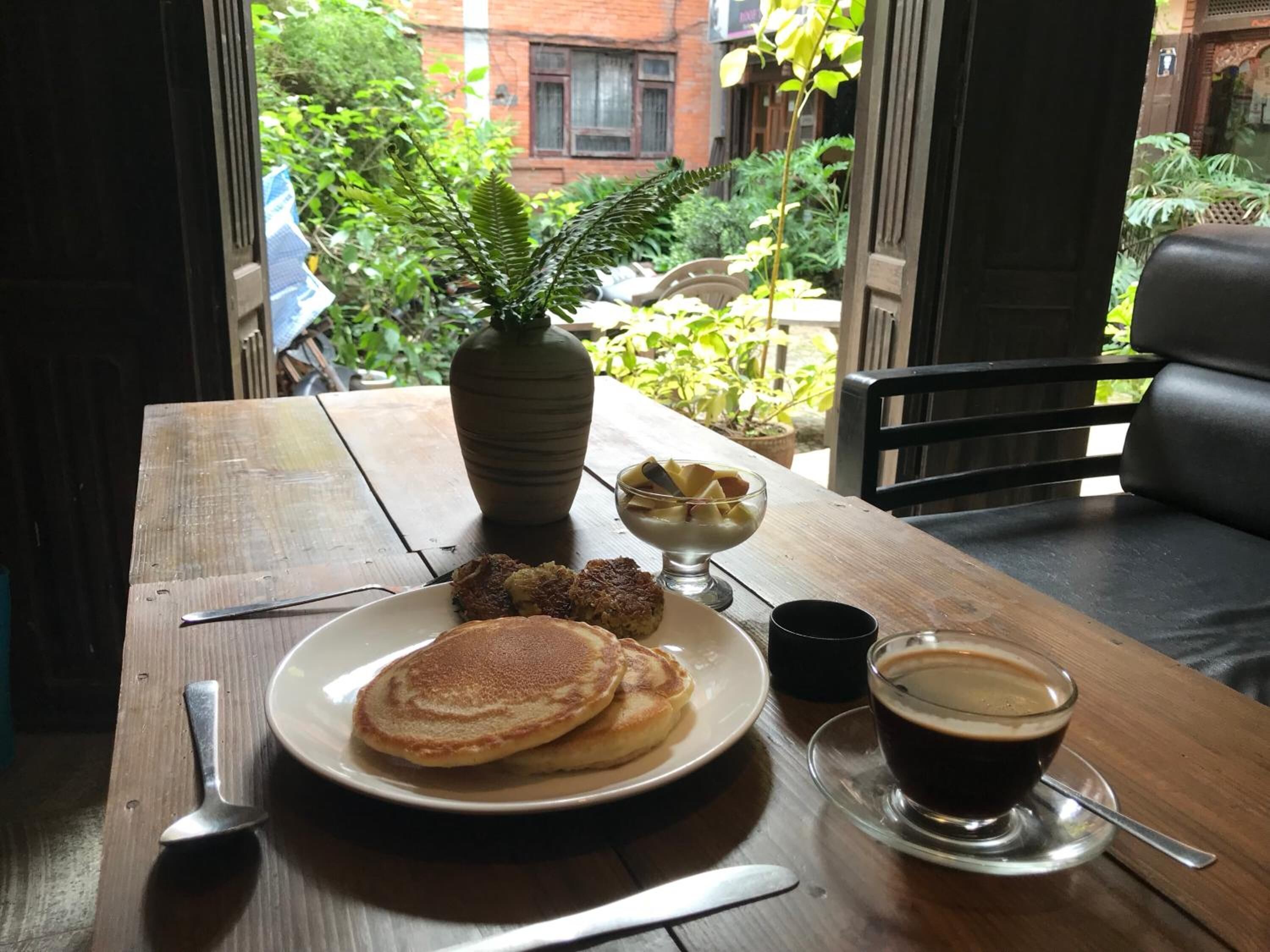 A breakfast setting with pancakes, a bowl of fruit, a cup of coffee, and a vase of flowers on a wooden table.