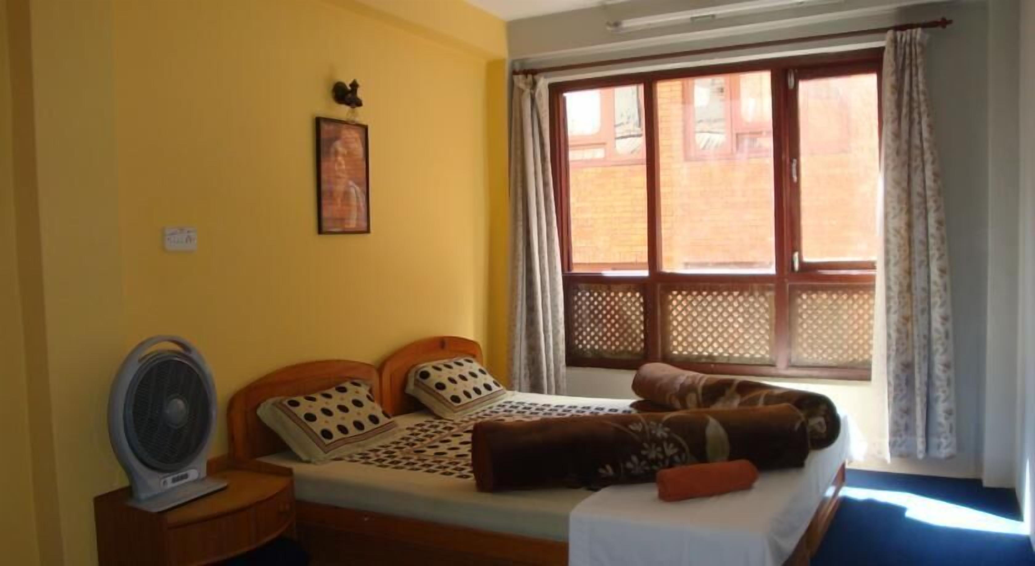 A room with a bed, a standing fan, a window with curtains, and a wall-mounted picture.
