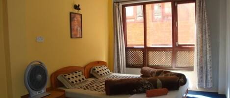 A room with a bed, a standing fan, a window with curtains, and a wall-mounted picture.