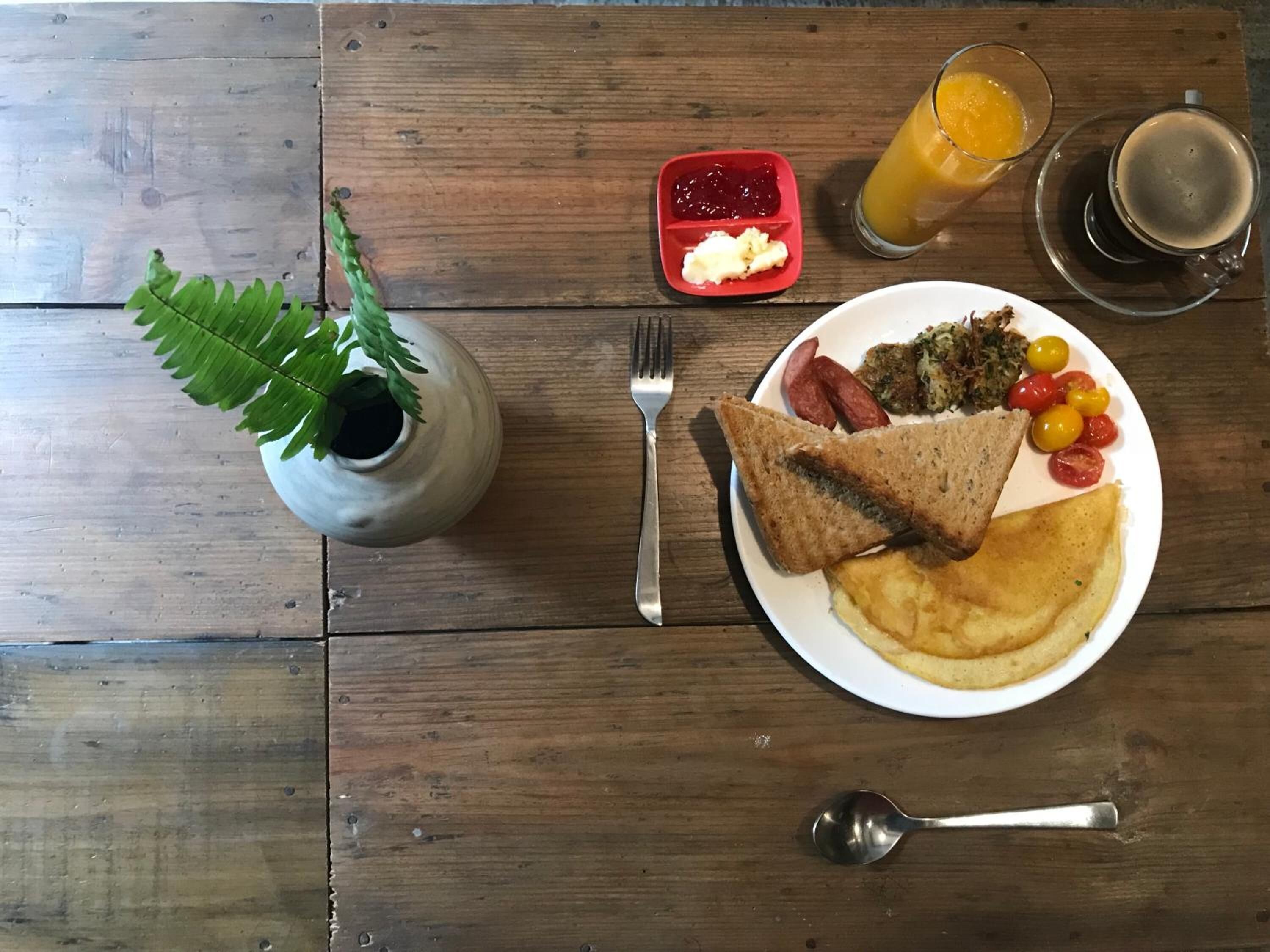 Une table en bois sur laquelle se trouvent une assiette de petit-déjeuner, un verre de jus d’orange, une tasse de café, un petit récipient rouge contenant de la confiture et une plante en pot.