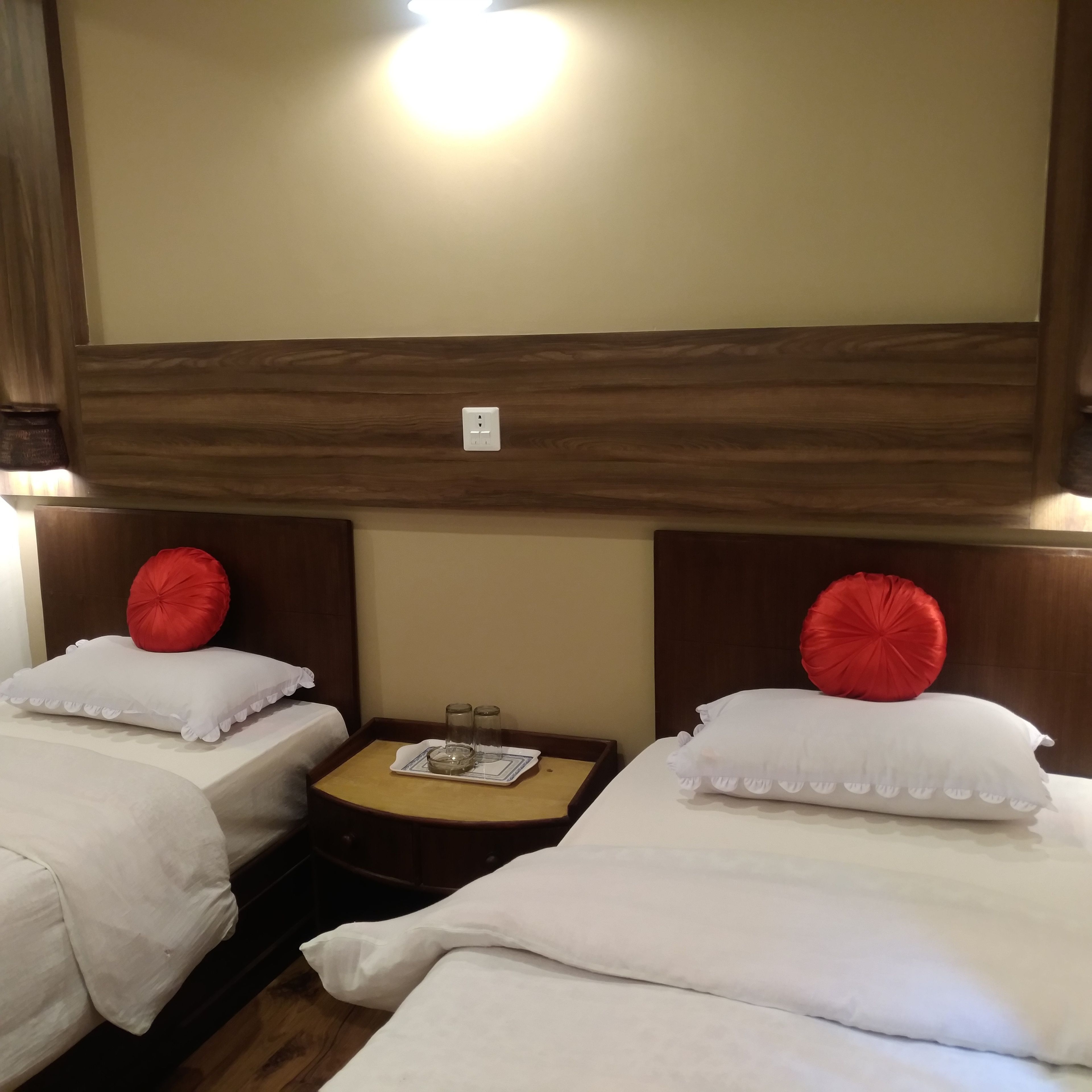 Une chambre d’hôtel avec deux lits, chacun doté de draps blancs et d’oreillers rouges, une petite table de chevet en bois avec un plateau, et un luminaire fixé au mur.