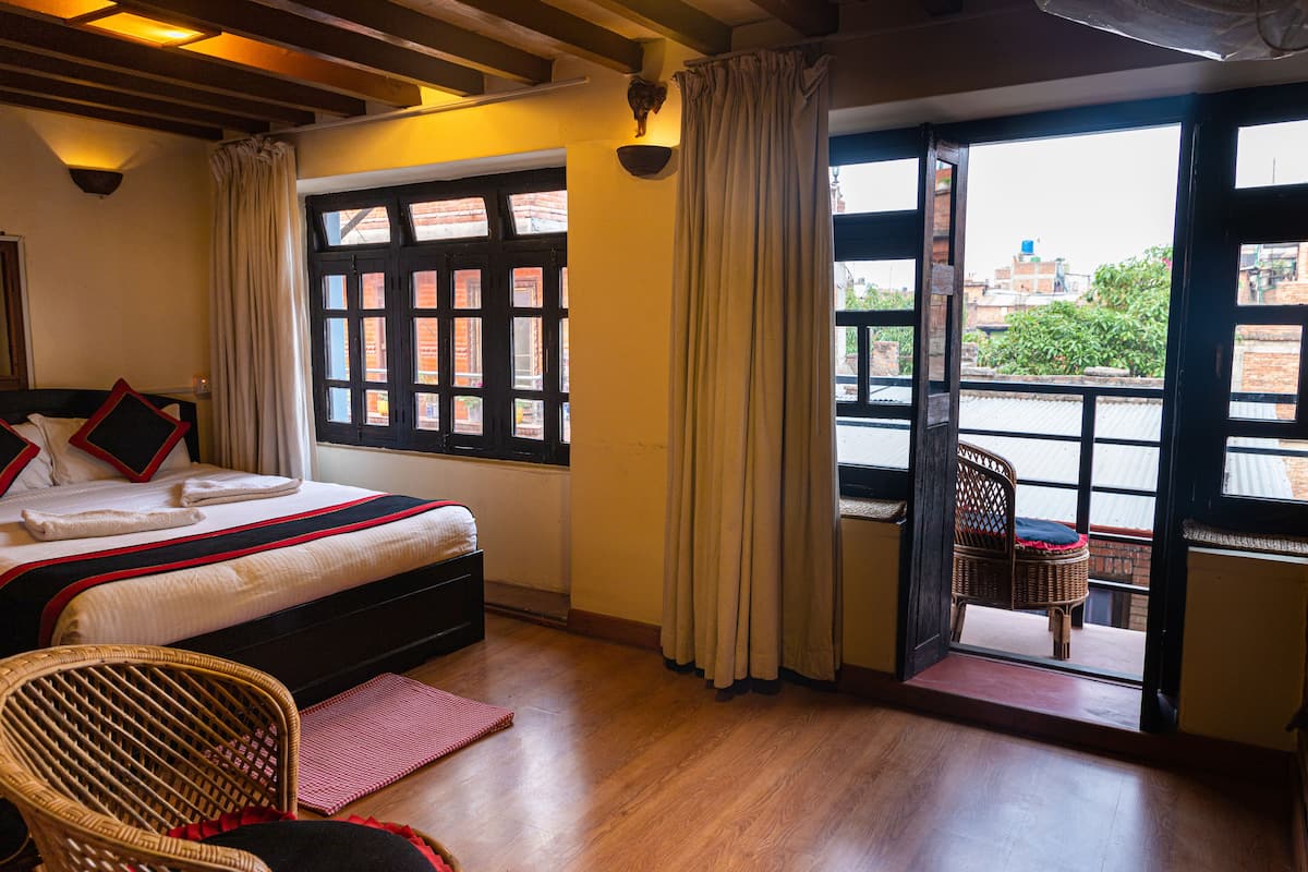 Een hotelkamer met een bed, een rieten stoel, een balkon met uitzicht op gebouwen en een raam met gordijnen.