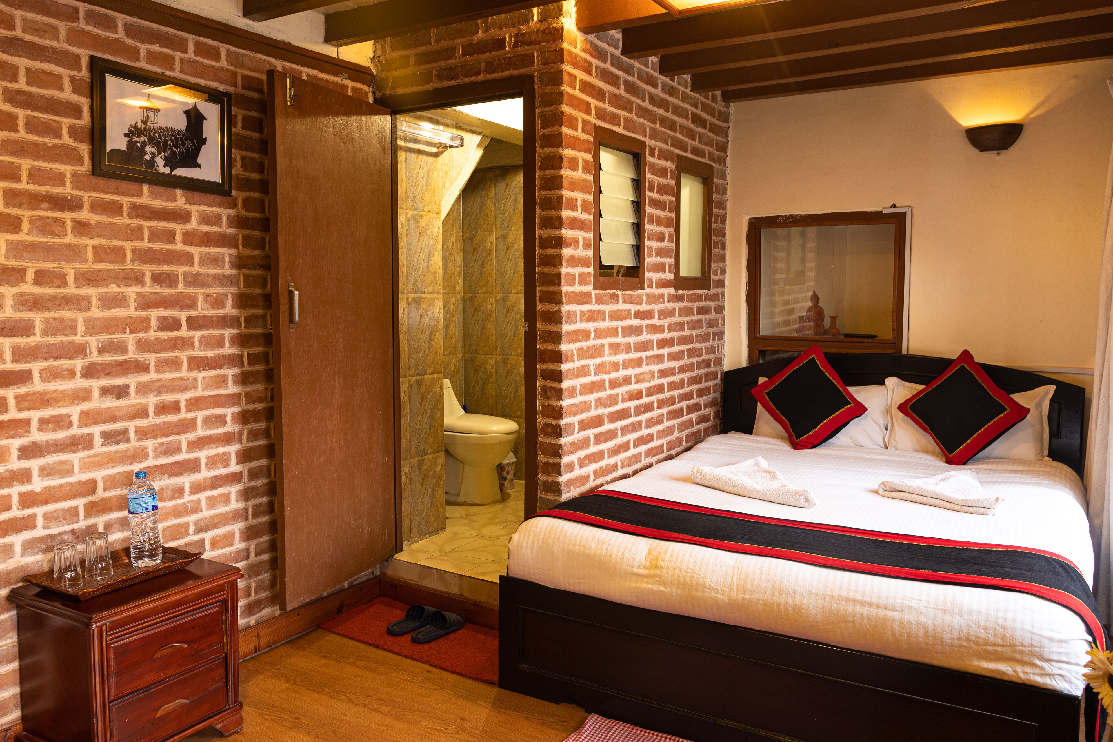 A hotel room with a brick wall, a wooden bed with red and black pillows, a wooden nightstand with a water bottle and glasses, and a wooden floor.