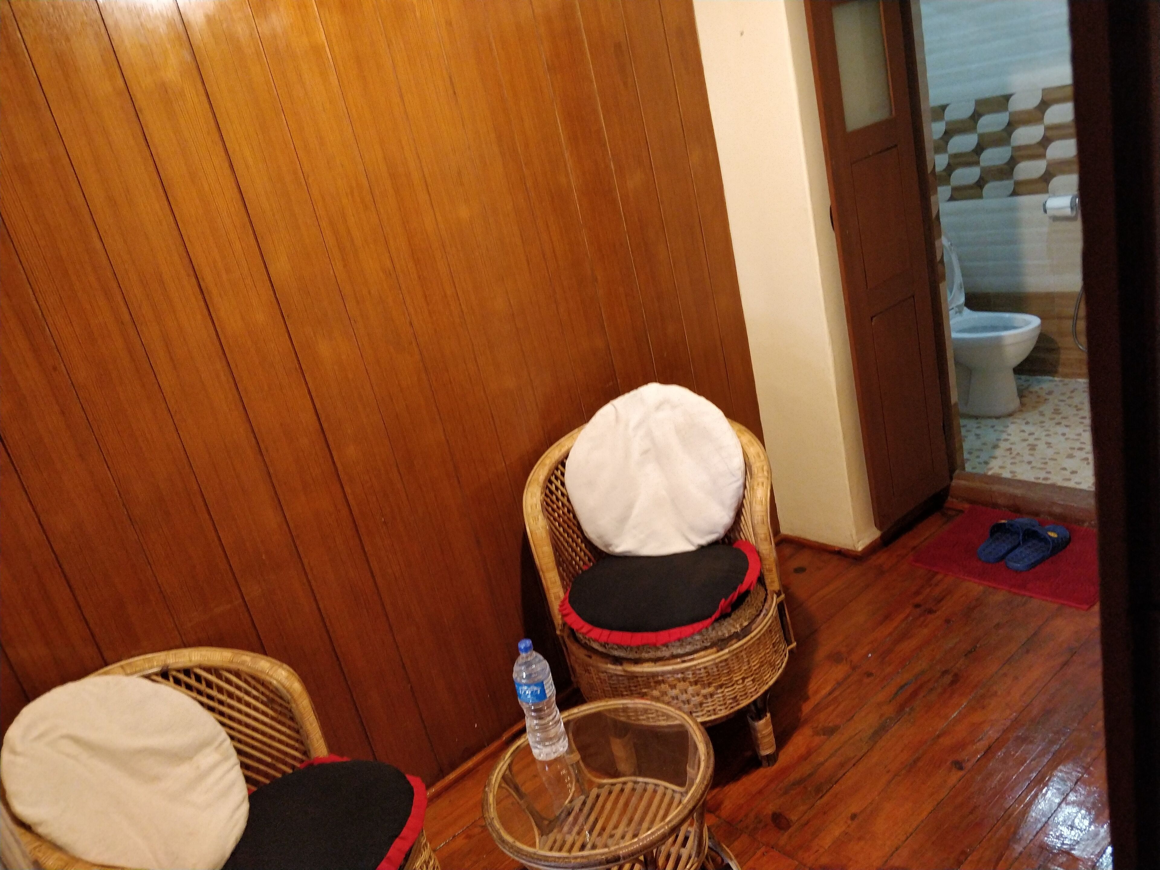 Une pièce avec un plancher en bois, une chaise en osier avec un coussin blanc, une petite table ronde avec une bouteille d’eau, et un tapis rouge près de la porte.