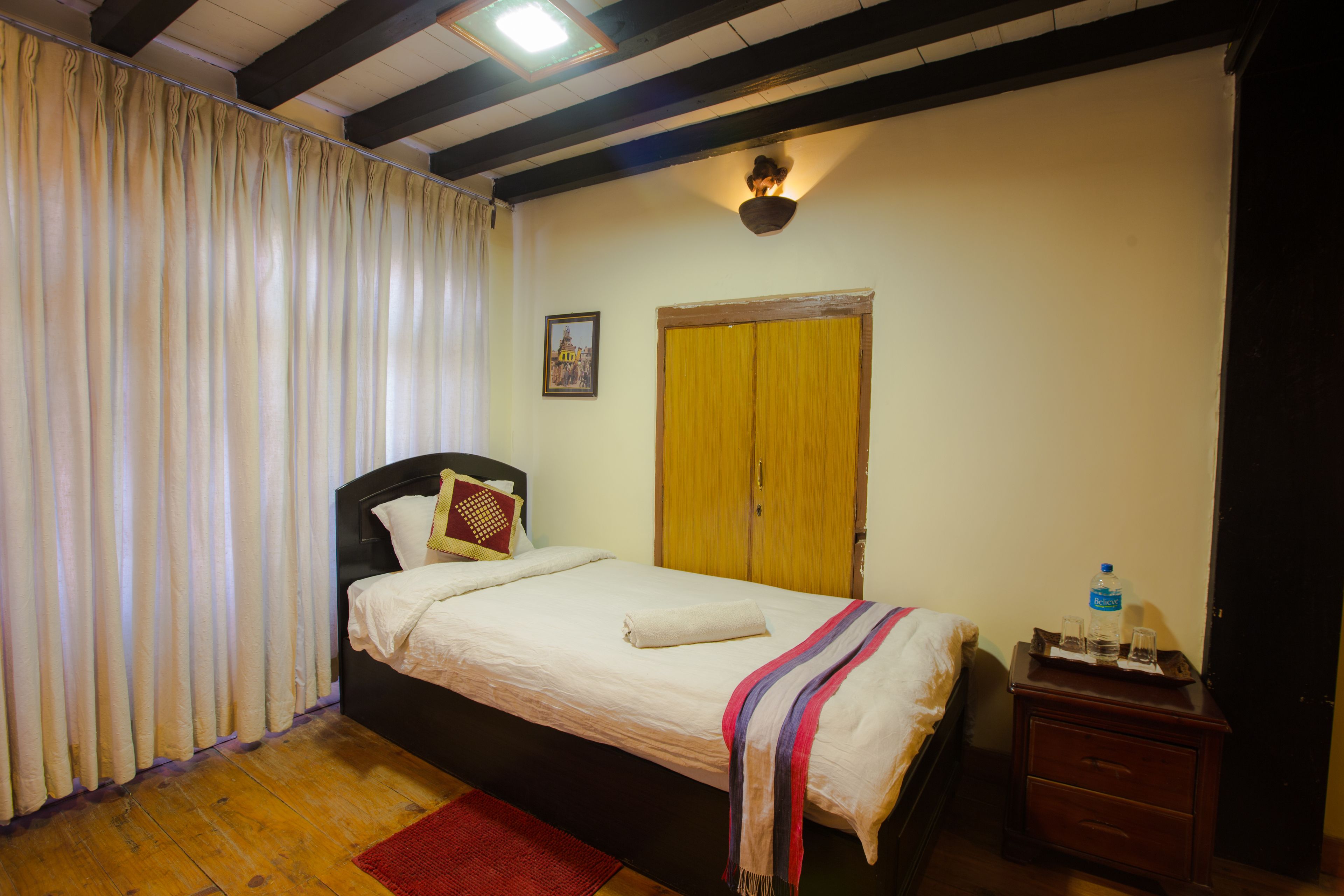Un dormitorio con cama, mesita de noche, piso de madera, alfombra roja, ventana con cortinas, lámpara de pared y puerta.