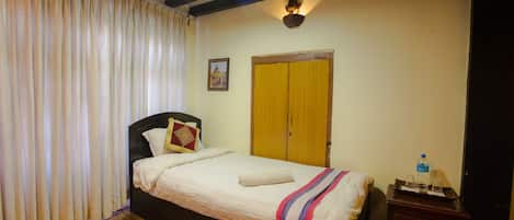 Un dormitorio con cama, mesita de noche, piso de madera, alfombra roja, ventana con cortinas, lámpara de pared y puerta.