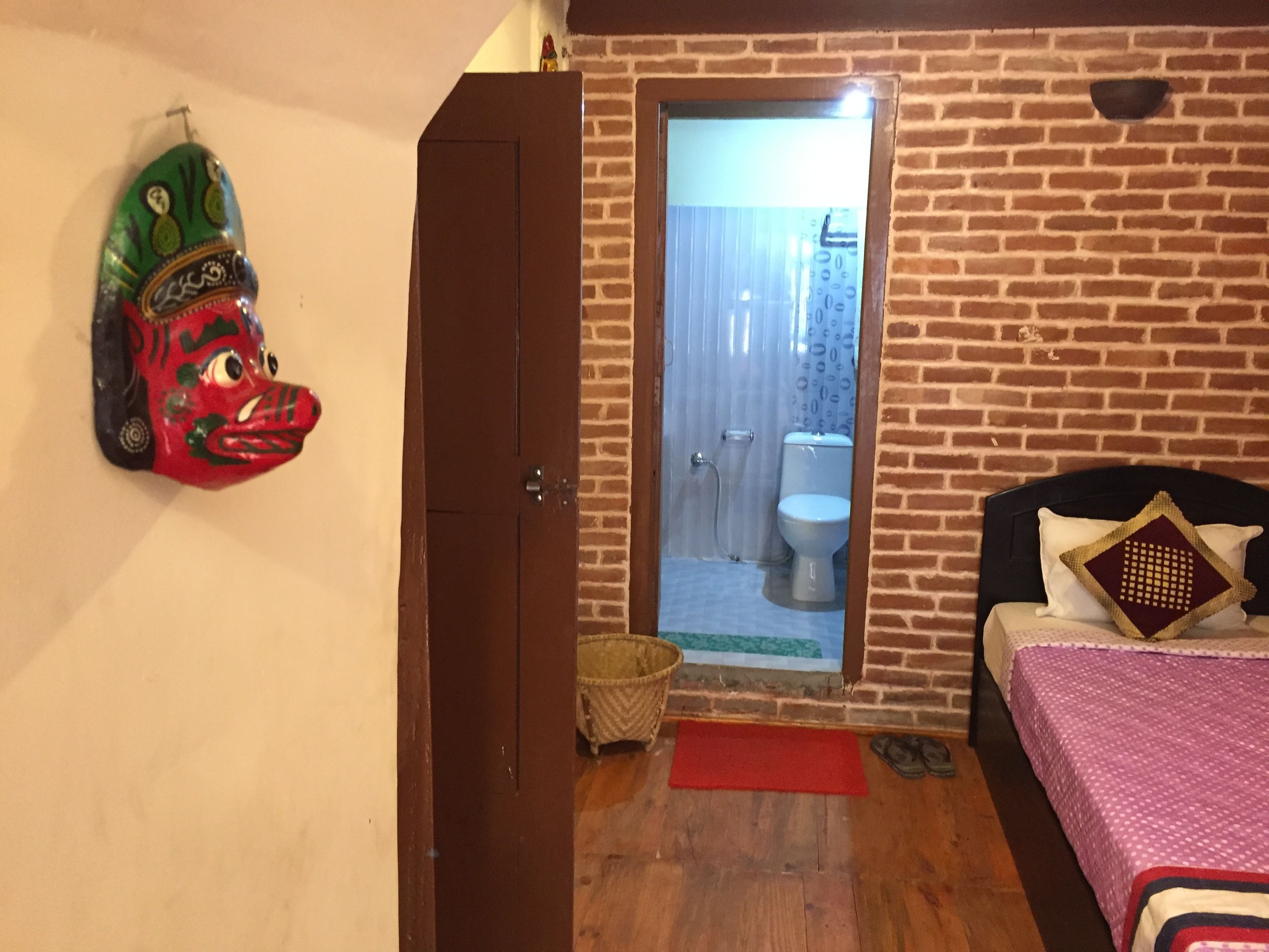 Une pièce avec une porte en bois donnant sur un balcon, un lit recouvert d’une courtepointe à motifs et un masque accroché au mur.