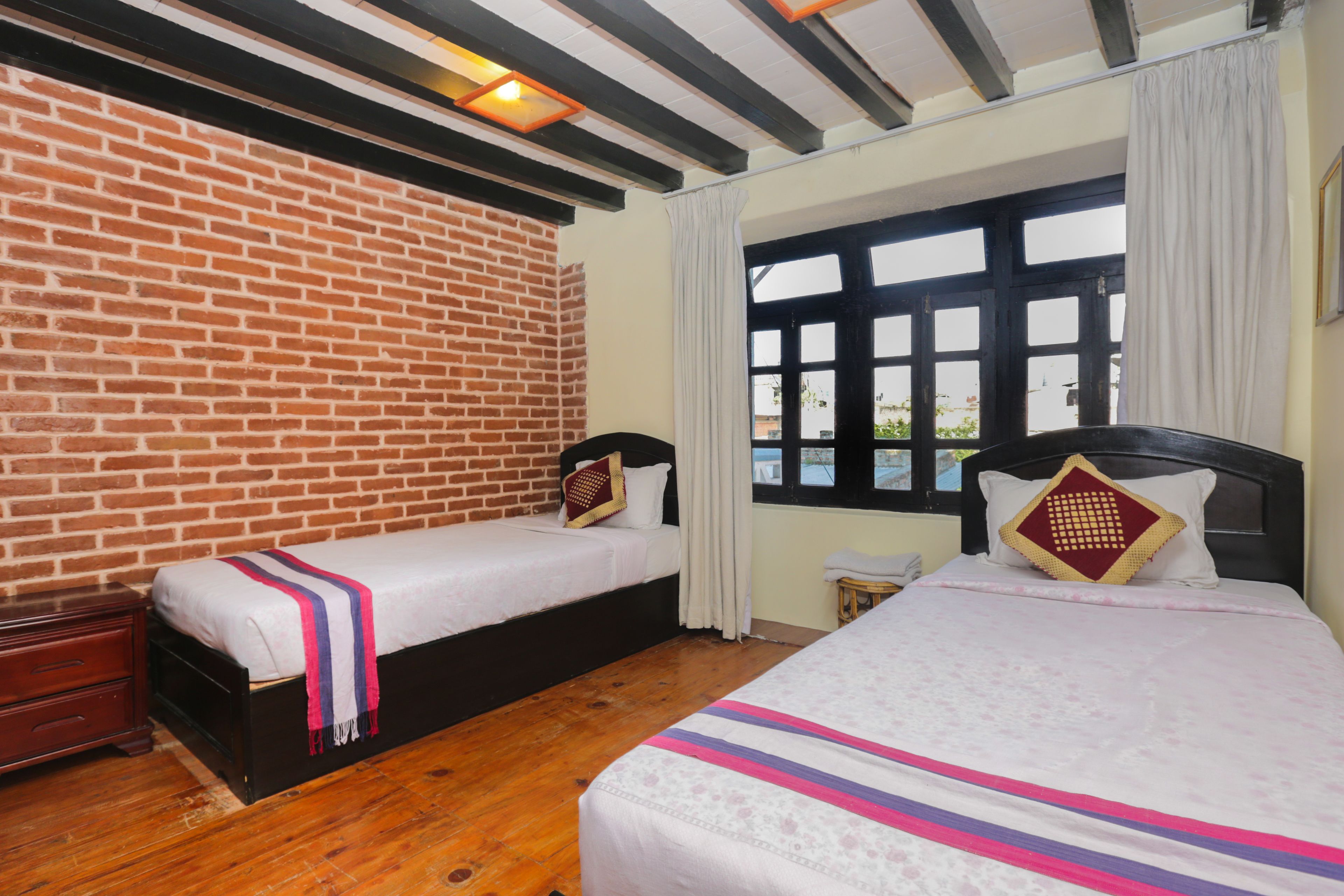 A room with two beds, a brick wall, a window with curtains, and a small table.