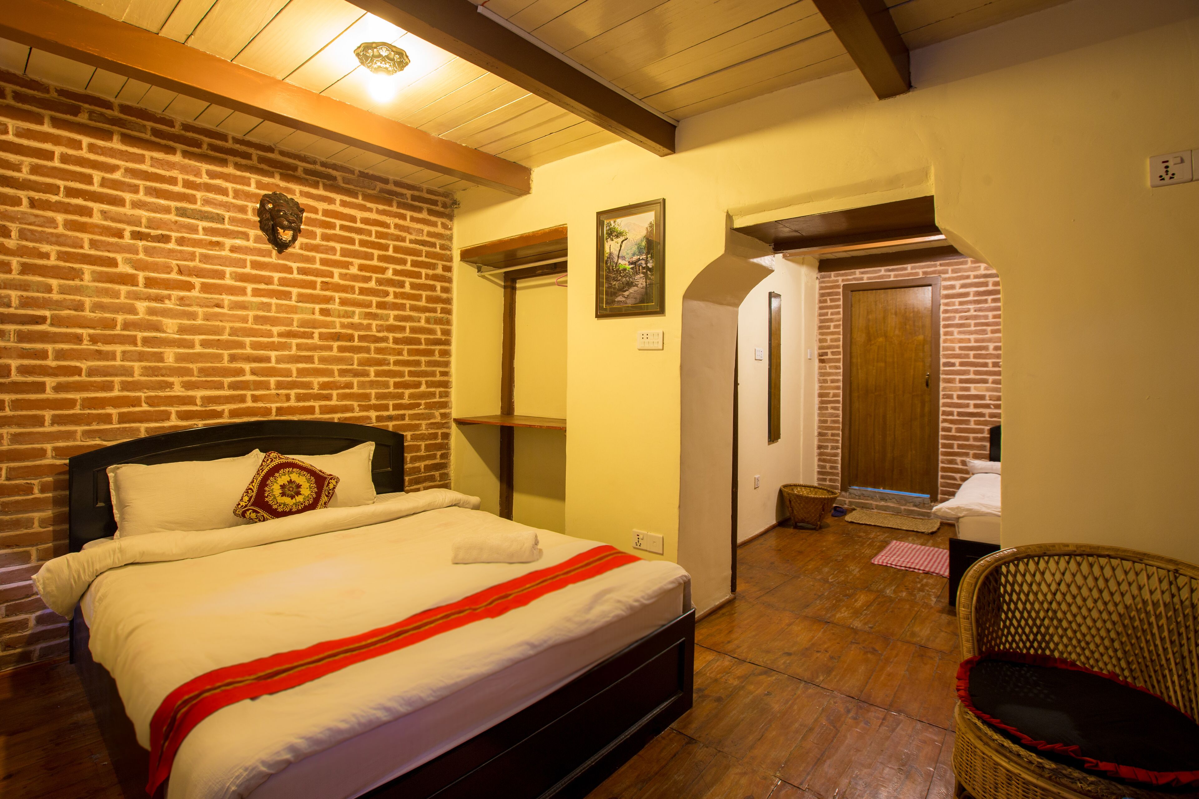 A bedroom with a brick wall, a wooden ceiling, a bed with white bedding, a wicker chair, and a door leading to another room.