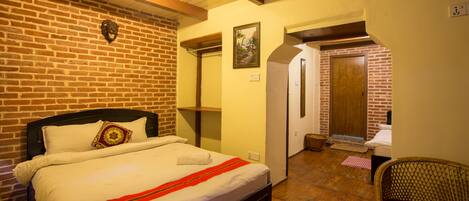 Un dormitorio con pared de ladrillo, techo de madera, cama con ropa de cama blanca, una silla de mimbre y una puerta que da a otra habitación.