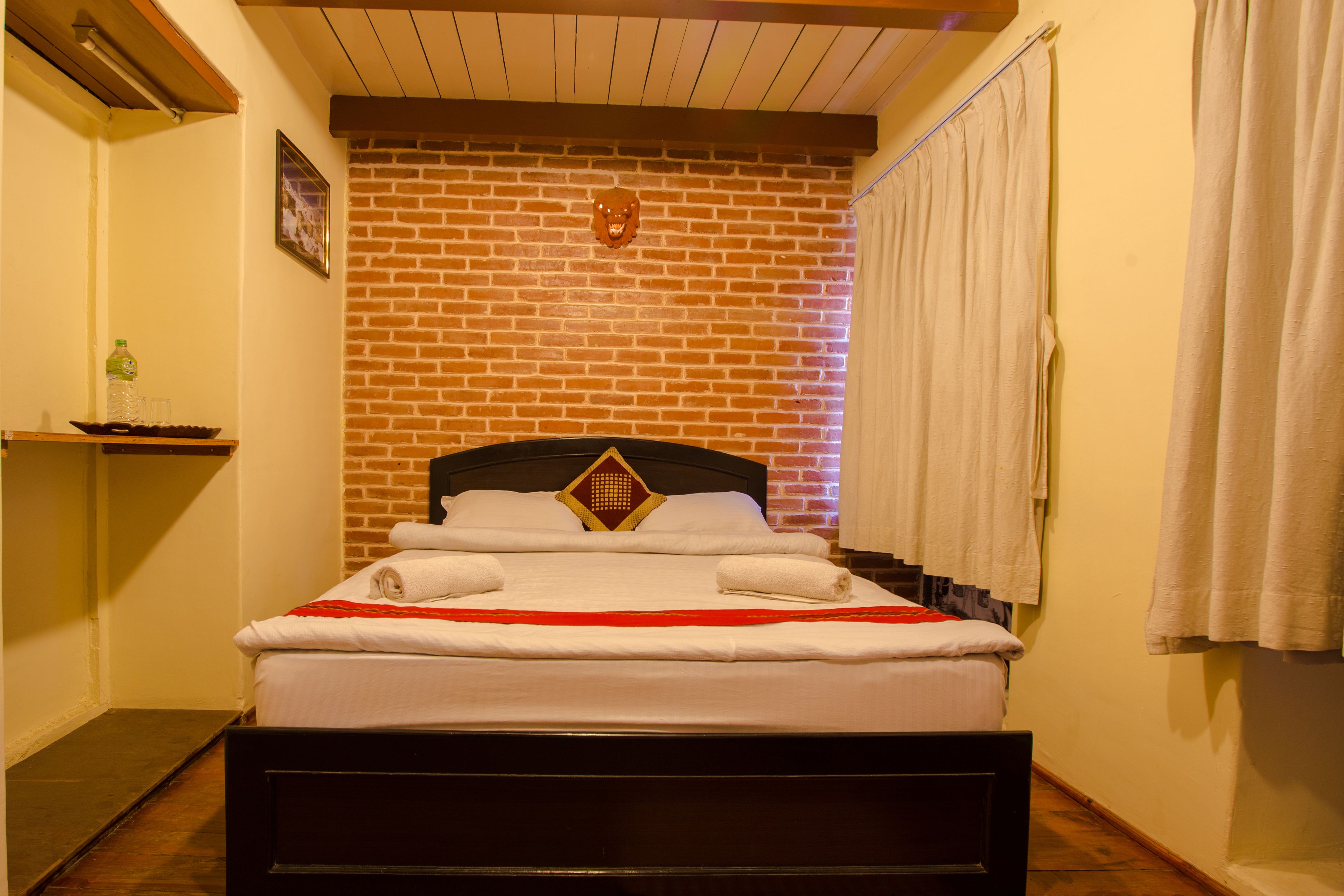 Uma cama bem arrumada com lençóis brancos e um toque de vermelho, parede de tijolos ao fundo e cabeceira de madeira.