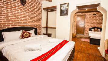 A hotel room with a brick wall, a bed with white bedding and a red accent, a wooden floor, and a wooden door leading to another room.