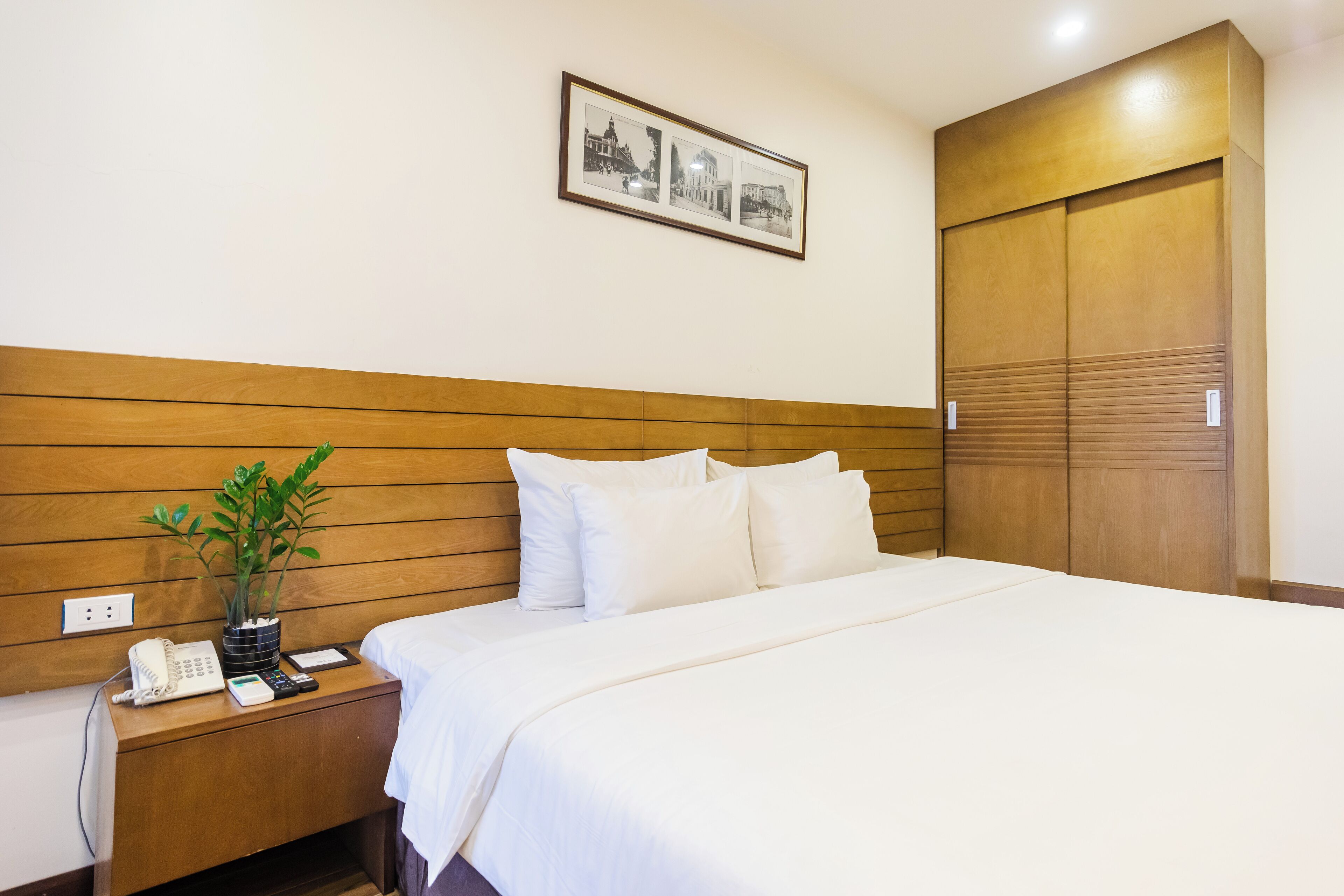 A hotel room with a wooden headboard, a bed with white bedding, a bedside table with a phone and a plant, and a framed picture on the wall.