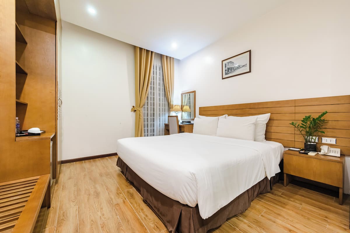 A hotel room with a large bed, wooden headboard, a bedside table with a lamp, a mirror, and a small plant.