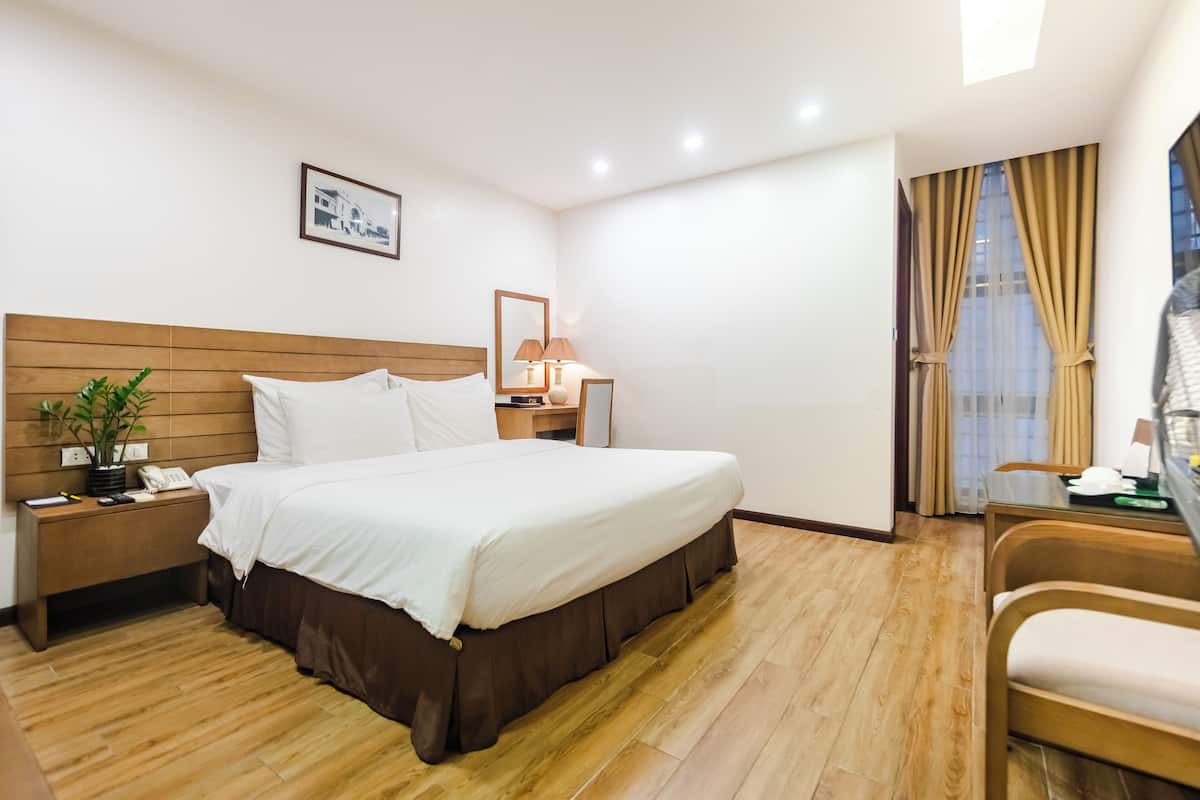 A hotel room with a wooden headboard bed, a bedside table with a lamp, a small desk with a chair, and a window with curtains.