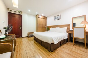 Habitación de hotel con piso de madera, cama con ropa blanca, escritorio con lámpara, armario de madera y ventilador de techo.