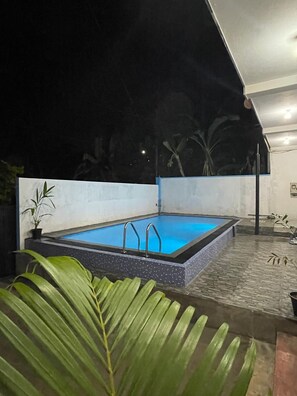 Una piscina rectangular con escaleras metálicas, rodeada por un suelo de baldosas y plantas en macetas.