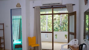 A room with a sliding glass door leading to a balcony, a yellow chair, a white chair, a wooden table with a kettle, and a small white bowl. - Feel Beach Apartments (Kalutara)