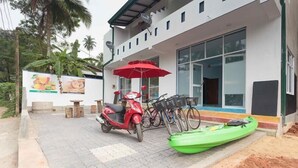 A modern building with a paved entrance, a red scooter, a green kayak, and bicycles parked outside. - Feel Beach Apartments (Kalutara)