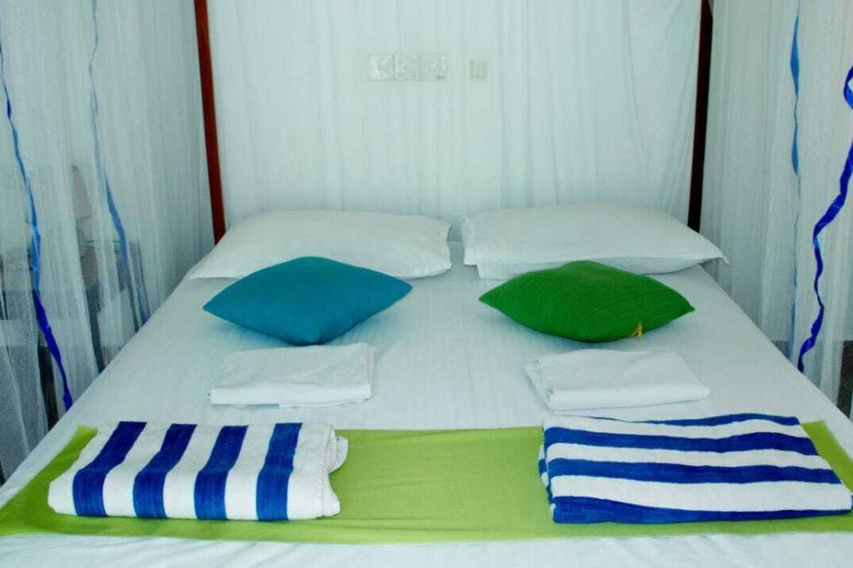 A single bed with white linens, two pillows (one blue, one green), and two striped towels (one blue, one green).
