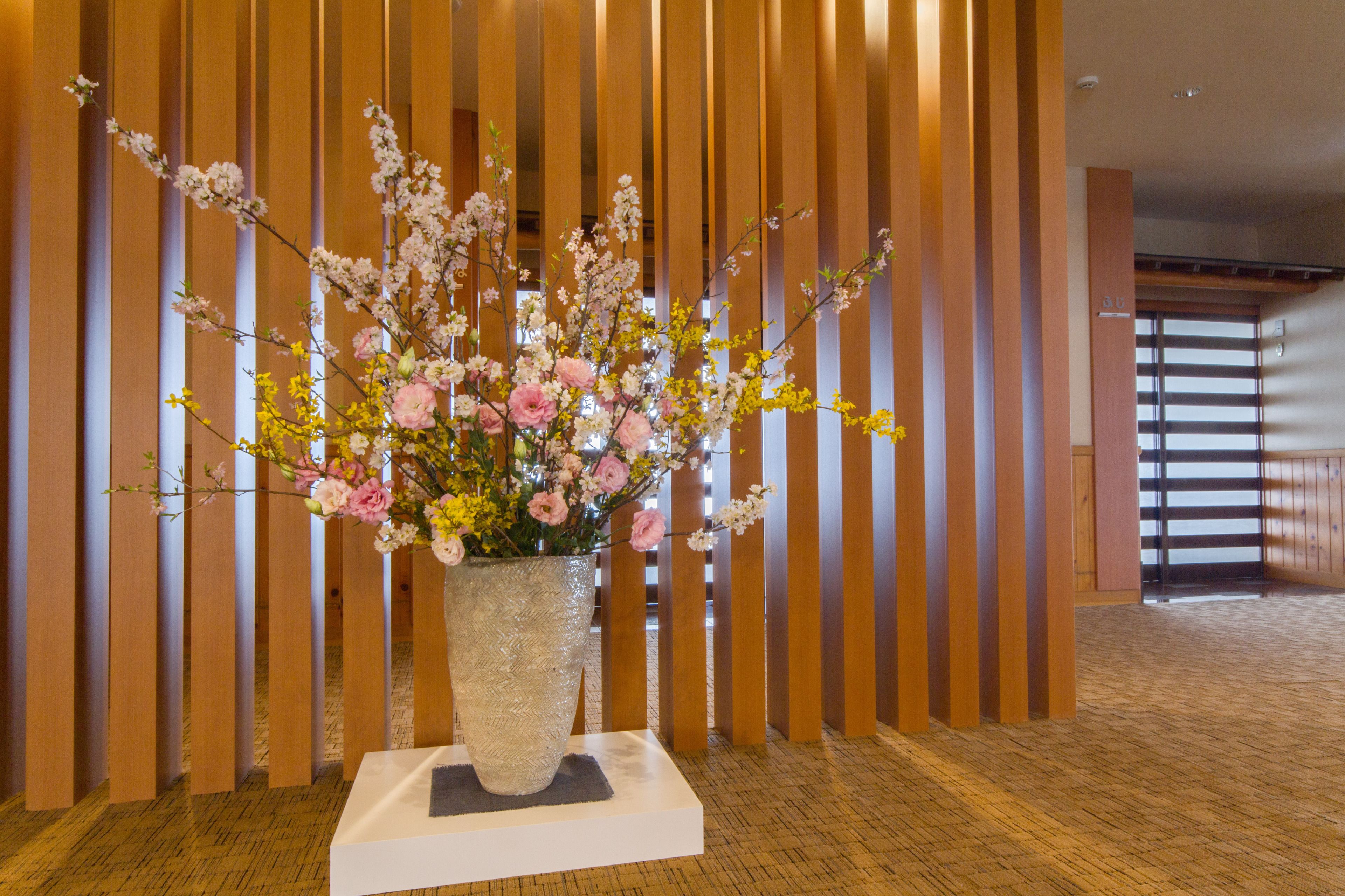 A large floral arrangement in a textured vase on a white pedestal in a hallway with wooden slats and a carpeted floor.