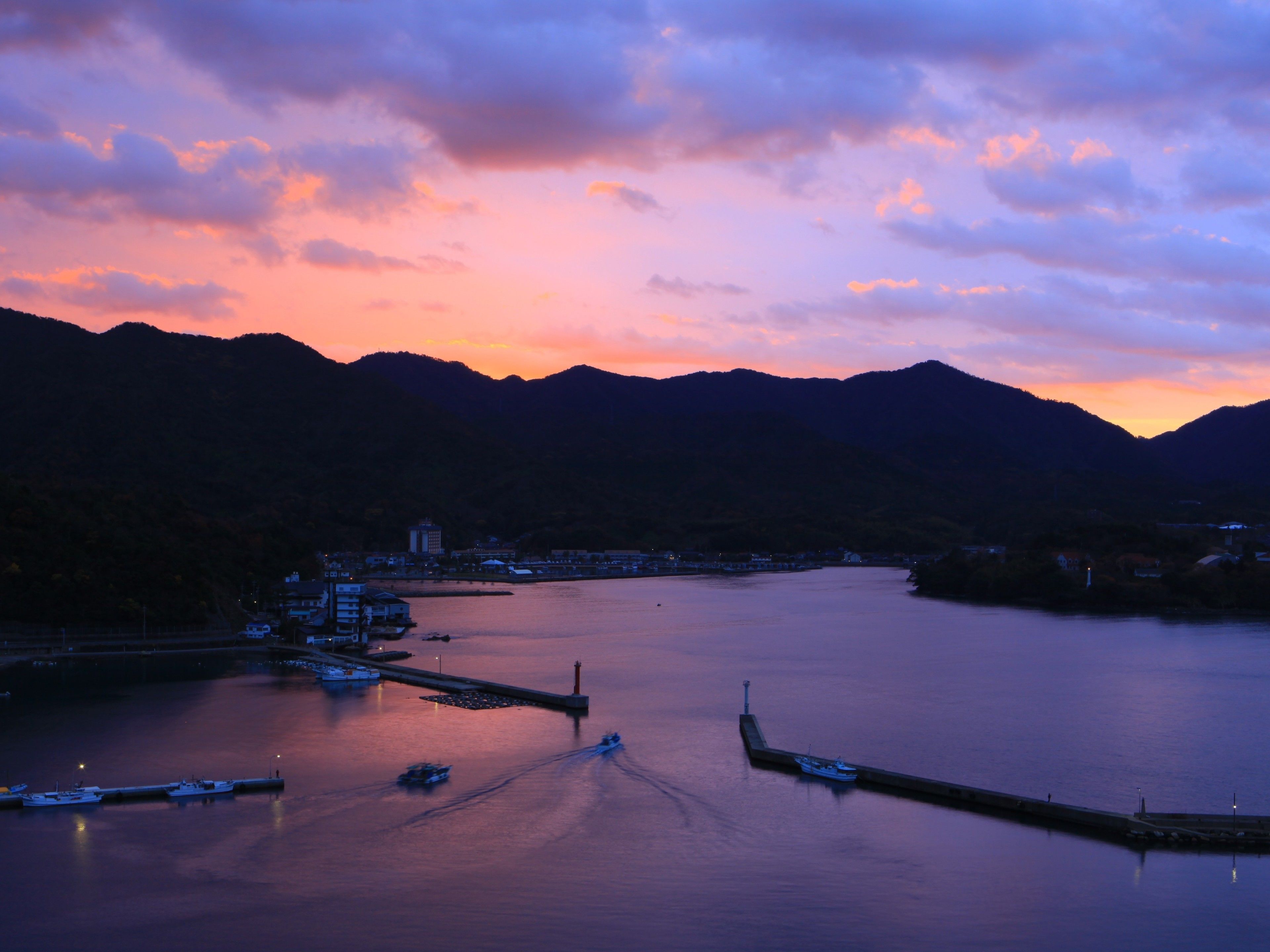 Un lago tranquilo con barcos amarrados en un muelle, rodeado de montañas y un pueblo al fondo.