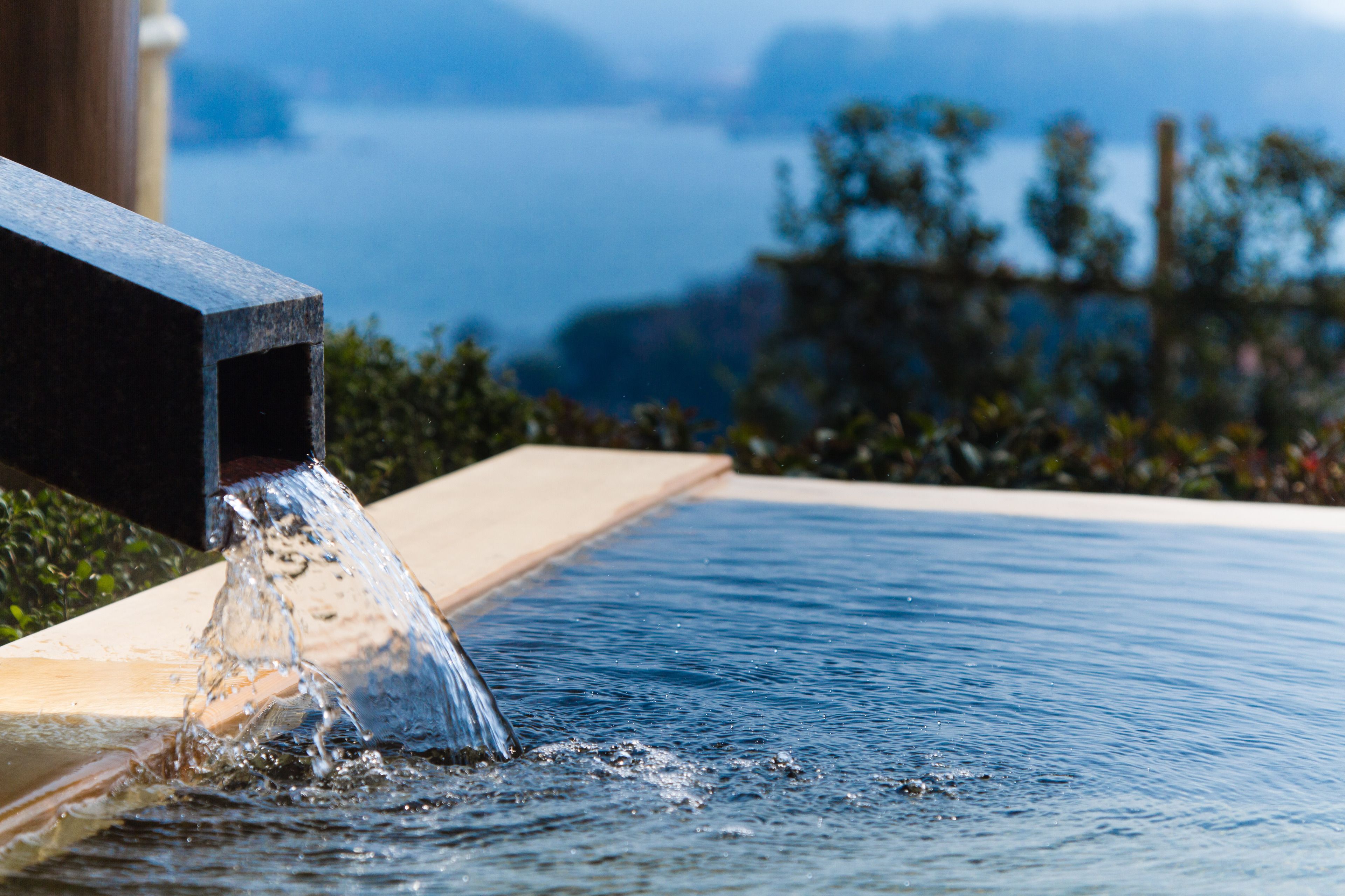 Una piscina con agua que cae desde una estructura, rodeada de vegetación y una vista distante.