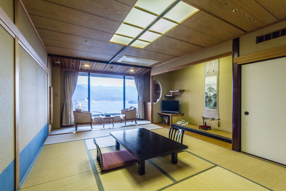 A traditional Japanese room with tatami flooring, a low table, chairs, a television, and a view of the sea.
