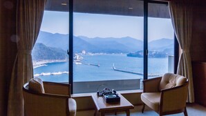 A room with a large window offering a view of a coastal area with mountains and a pier. There are two chairs and a small table with a stereo system. - Hagi Kanko Hotel (Hagi)