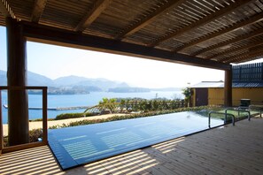 Piscina infinita con vistas a un lago y montañas, terraza de madera y zona cubierta con vistas.