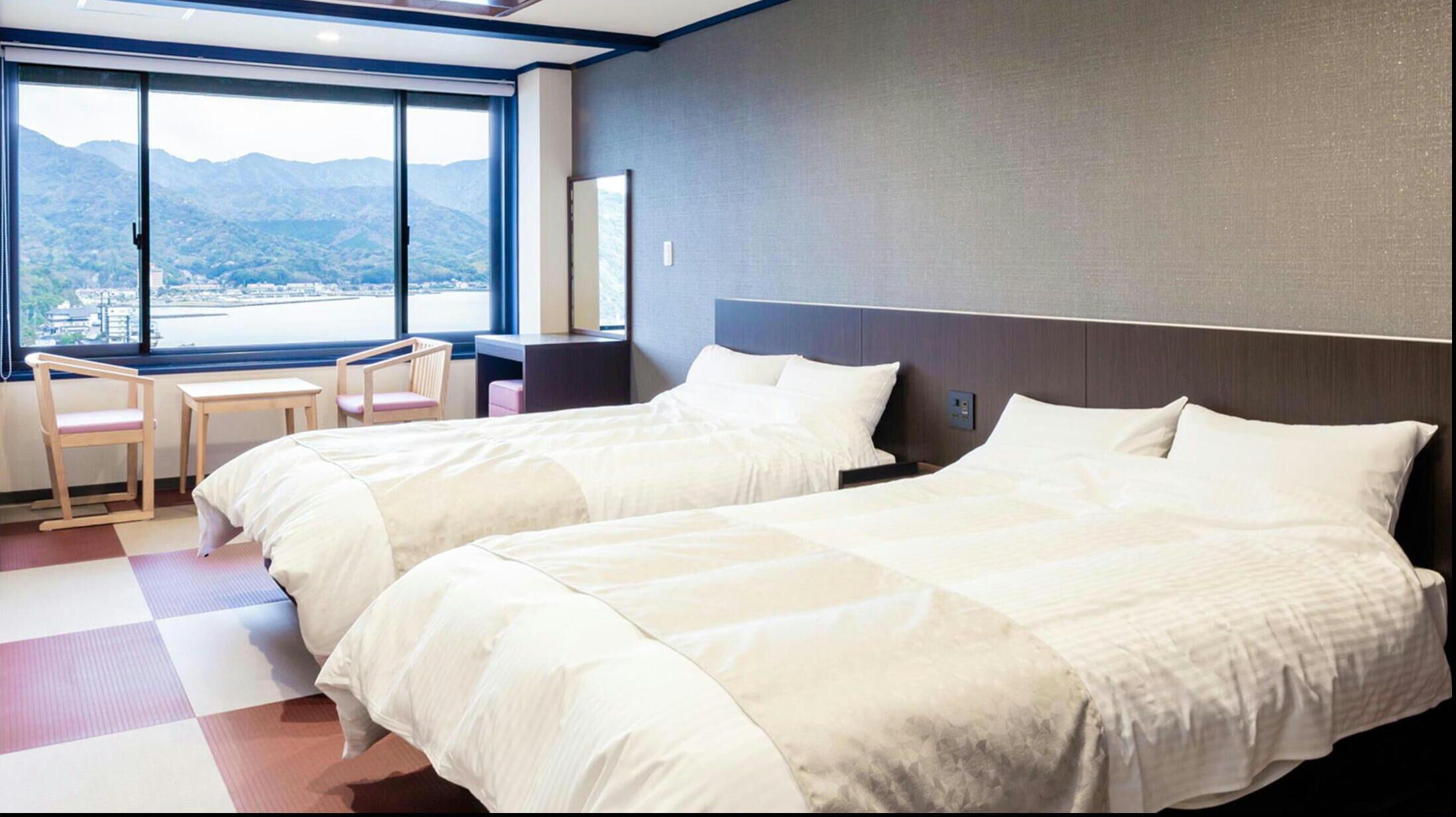 Habitación de hotel con dos camas, un escritorio, una silla y un ventanal con vista a la montaña.