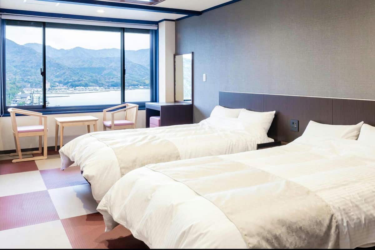 A hotel room with two beds, a desk, a chair, and a large window with a mountain view.