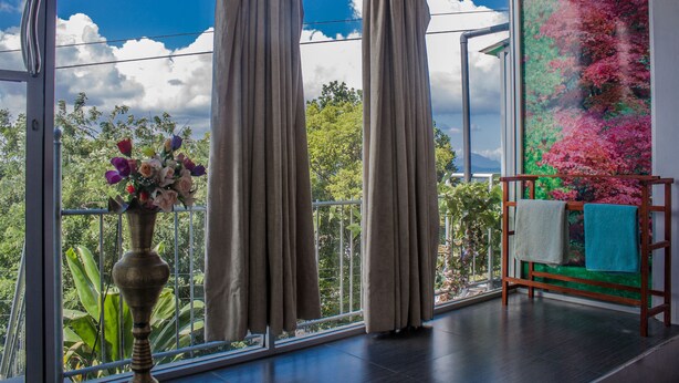 Una habitación con un ventanal grande, cortinas, un florero con flores y un perchero de madera con toallas.