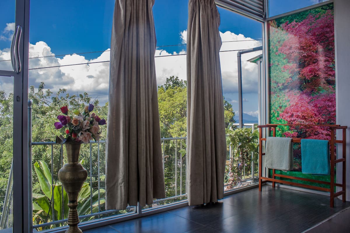 Ein Zimmer mit großem Fenster, Vorhängen, einer Vase mit Blumen und einem Holzregal mit Handtüchern.
