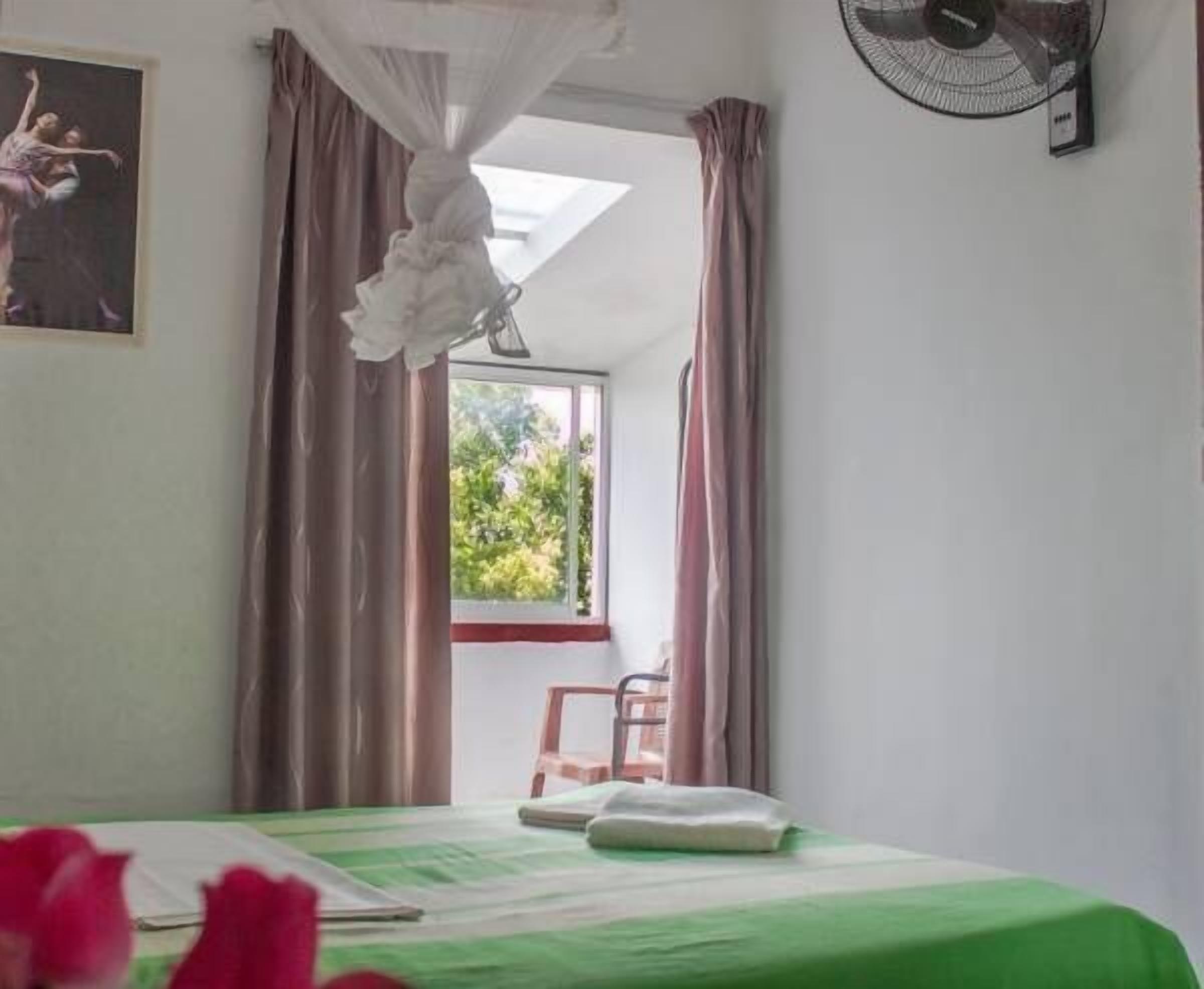 Une pièce comprenant un lit, une chaise, une fenêtre avec des rideaux et un ventilateur fixé au mur.