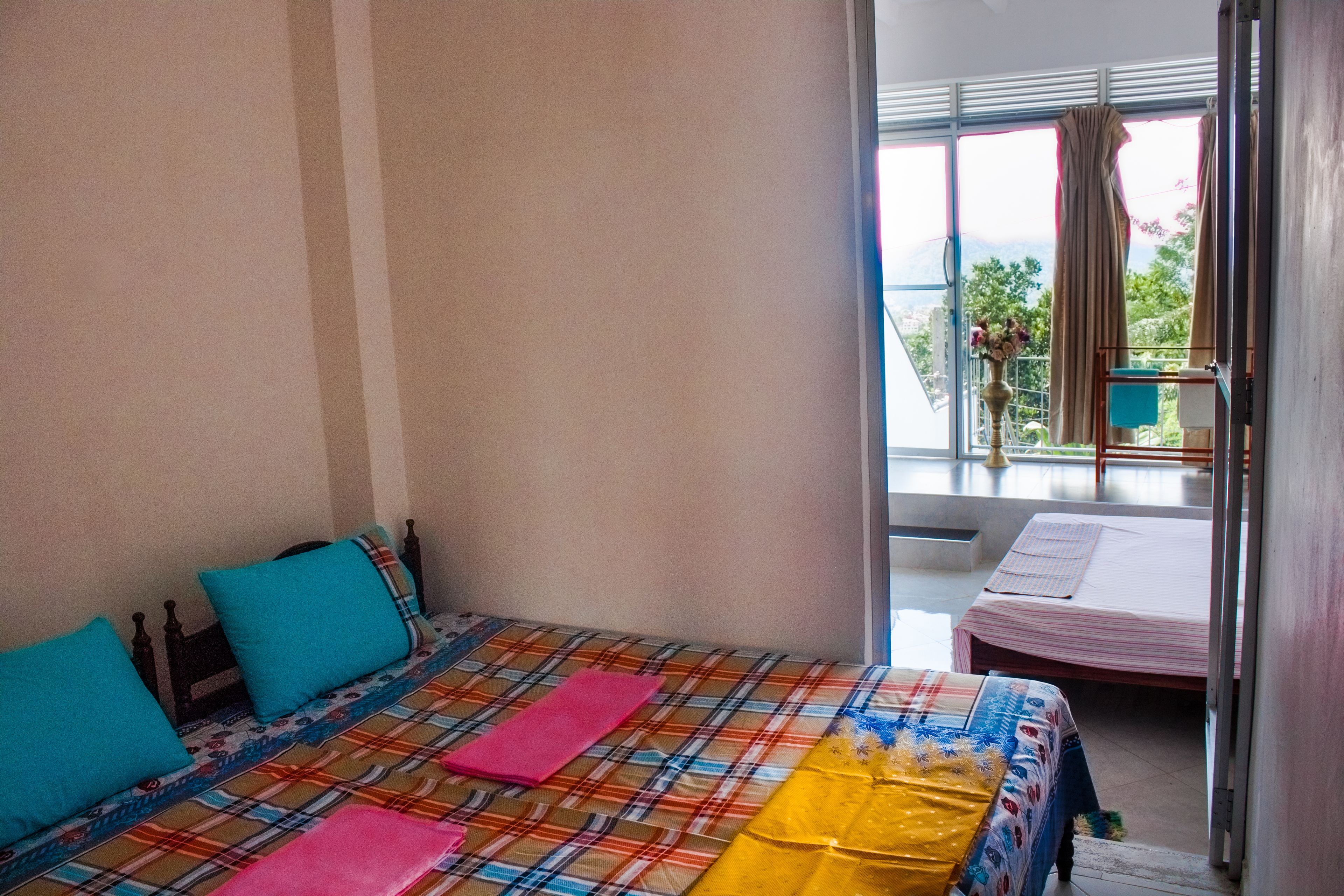 Una habitación con dos camas, una con una colcha escocesa de colores y la otra con una colcha blanca lisa. Hay una ventana con cortinas, y un pequeño balcón con una silla y una mesa.