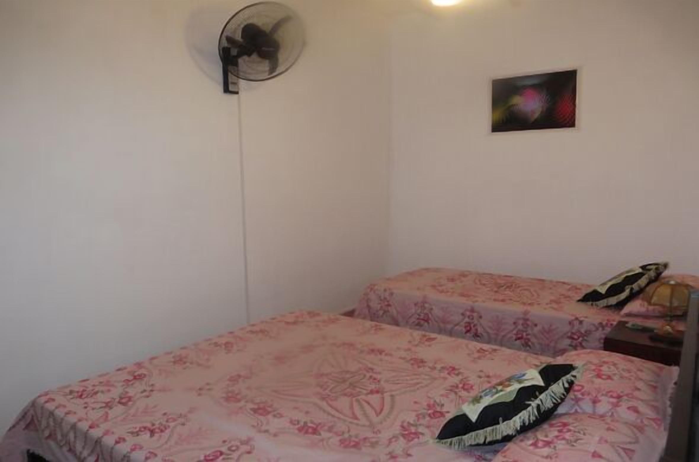 A small, simple room with two beds, each with floral bedding. There is a wall-mounted fan and a single piece of wall art.
