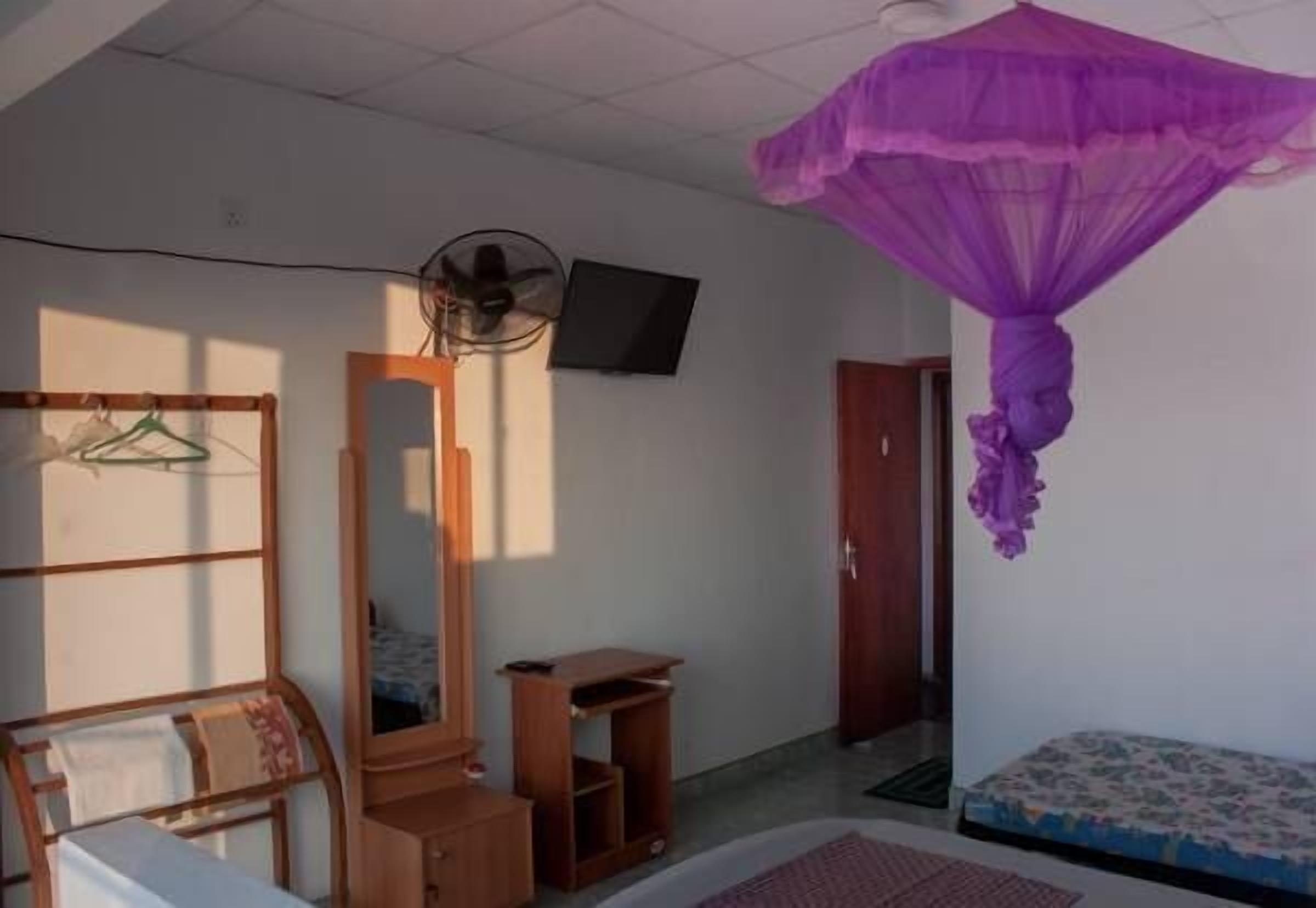 A bedroom with a bed, a wooden wardrobe, a mirror, a small table, a hanging clothes rack, and a purple canopy.