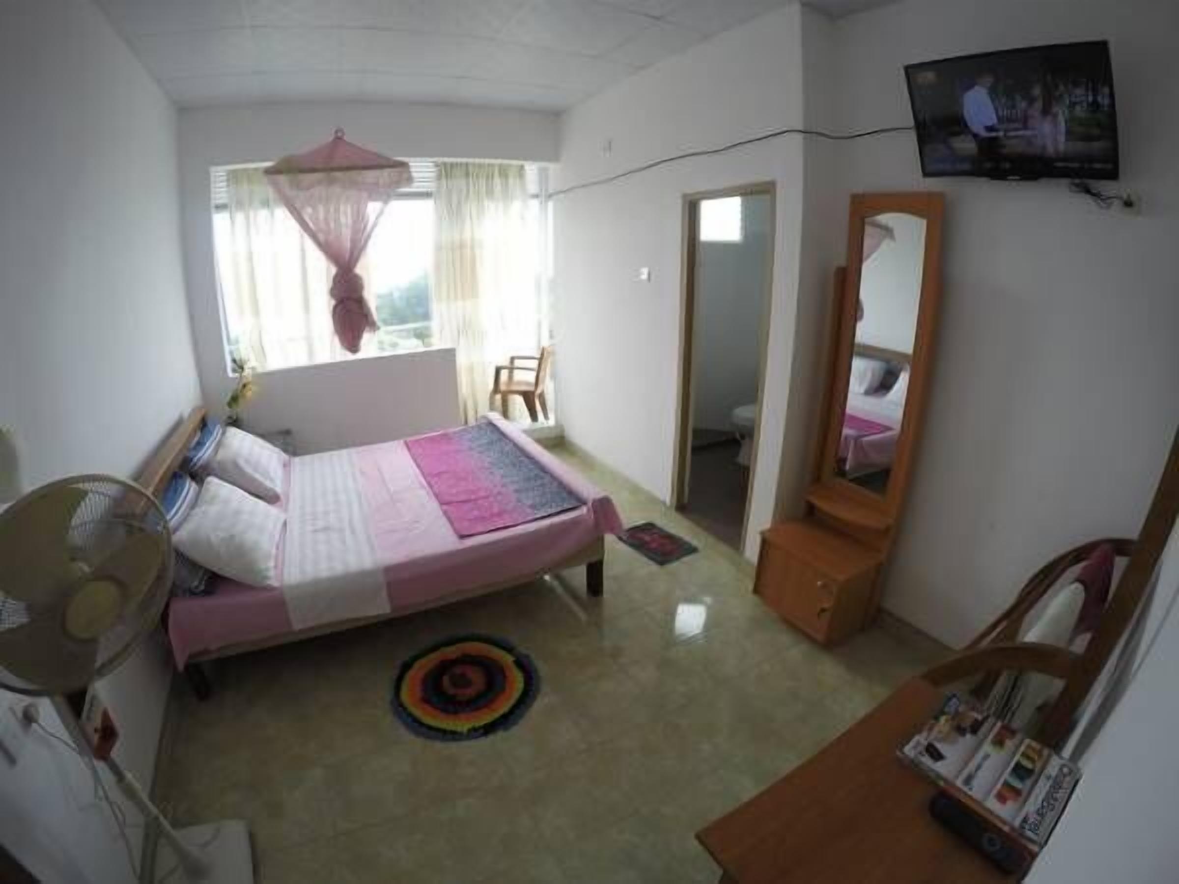 Un dormitorio con cama, mesita de noche, espejo, televisor y ventana con cortinas.