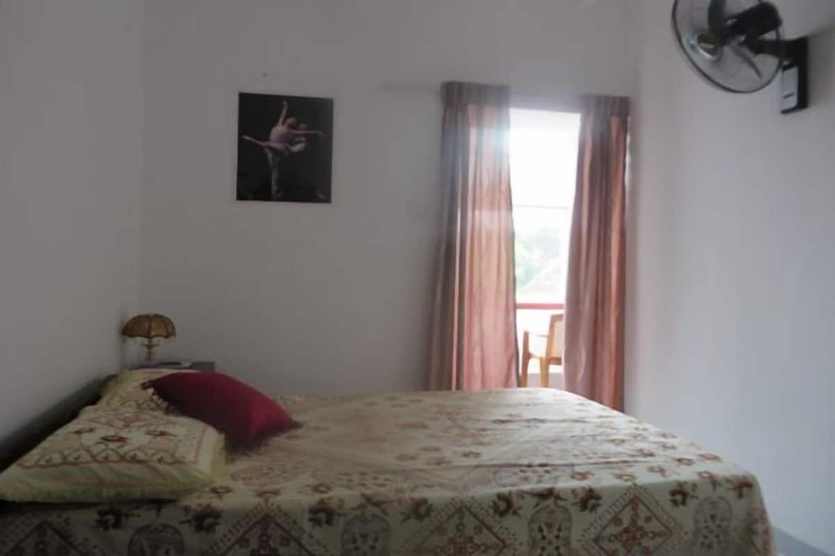 Ein Schlafzimmer mit Bett, Wandventilator, Fenster mit Vorhängen und einem Bild an der Wand.