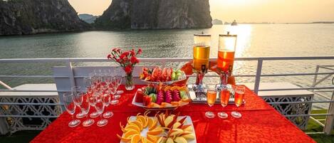 Une table dressée avec des fruits, des verres et un distributeur de boissons, donnant sur une étendue d’eau bordée de formations rocheuses.